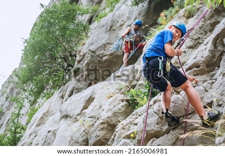 Smiling teen boy in protective helmet abseiling from cliff rock wall using rope, Belay device and climbing harness with father in Paklenica Park Croatia. Active extreme sports time spending concept Royalty-Free Stock Photo #2165006981