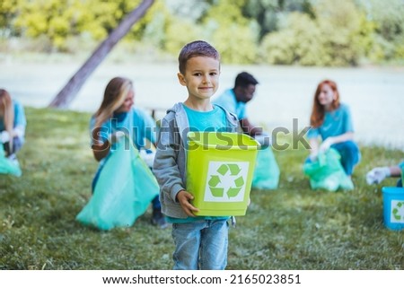 Portrait of a little schoolboy with short hair holding a green crate with a white recycling logo on it and looking to camera smiling in city park