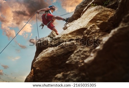 Muscular climber man in protective helmet abseiling from cliff rock wall using rope Belay device and climbing harness on evening sunset sky background. Active extreme sports time spending concept. Royalty-Free Stock Photo #2165035305