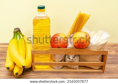 Donation food box, volunteering help,humanitarian aid full of canned,sunflower oil, pasta,cereal,grain and apples,banana,fruits.Delivery wooden box for charity on yellow, wooden background still life.