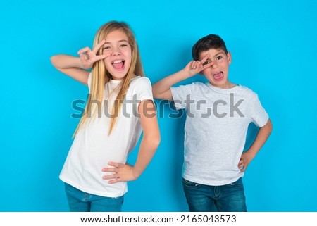 two kids boy and girl standing over blue background making v-sign near eyes. Leisure, coquettish, celebration, and flirt concept.
