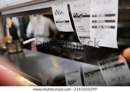 "check on" ticket orders on the pass of a professional kitchen with chefs cooking in the background. "on deck" menu items organised by waiters in order of priority for the customers.Grill, hob, oven,  Royalty-Free Stock Photo #2165050299