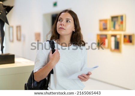 Focused girl visitor with a booklet in her hands examines an exhibit standing in an art gallery Royalty-Free Stock Photo #2165065443