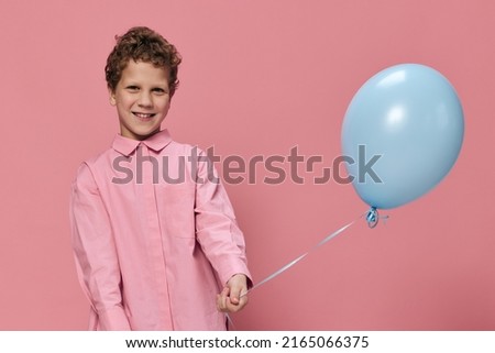 portrait of a funny, very beautiful, curly-haired boy standing in pink clothes on a pink background with a blue balloon in his hand