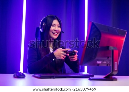 Playing video game. Young asian pretty woman sitting on chair holding joystick in living room. Happy female Professional Streamer wearing headphone playing game in dark room neon light.