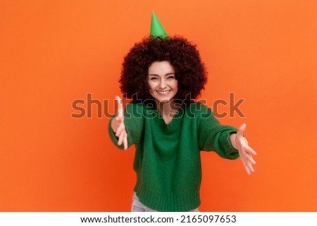Happy satisfied woman with Afro hairstyle wearing green casual style sweater and cone, celebrating birthday party, inviting guests. Indoor studio shot isolated on orange background.