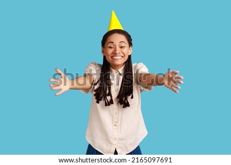 Come into my arms. Friendly woman with black dreadlocks and party cone on head giving free hugs with outstretched hands, welcoming inviting to embrace. Indoor studio shot isolated on blue background.