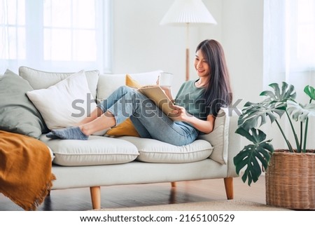 Young asian woman sitting on modern sofa in front of window relaxing in her living room reading book and drinking coffee or tea at home
