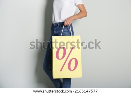 Woman holding paper shopping bag with percent sign on light grey background, closeup. Discount concept