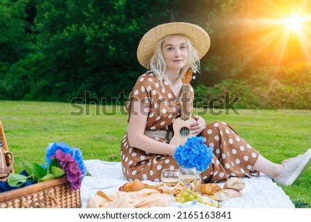 Girl in polka dot dress and hat sitting on white knit picnic blanket plays ukulele and drinking wine. Summer picnic on sunny day with bread, fruit, bouquet hydrangea flowers. Selective focus