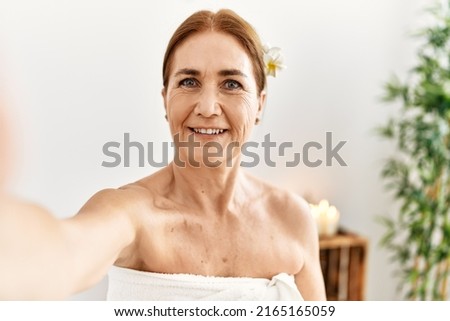 Middle age caucasian woman wearing towel make selfie by the camera sitting on massage board at beauty center