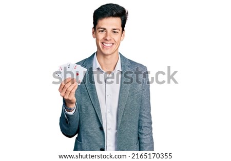 Young hispanic business man playing poker holding cards looking positive and happy standing and smiling with a confident smile showing teeth 