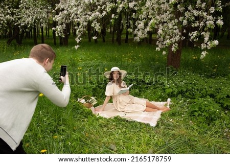 pregnant woman reading a book at a picnic. Husband takes a picture of his pregnant wife on the phone