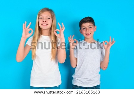 Glad two kids boy and girl standing over blue background shows ok sign with both hands as expresses approval, has cheerful expression, being optimistic.