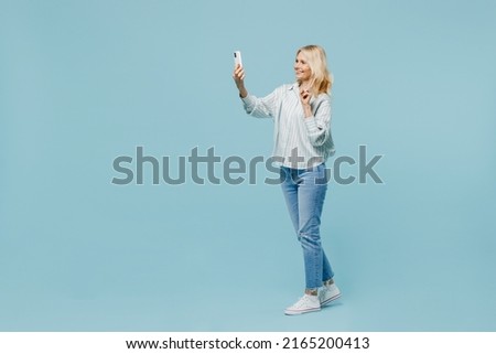 Full body elderly woman 50s wearing casual striped shirt doing selfie shot on mobile cell phone post photo on social network showv-sign gesture isolated on plain pastel light blue color background.