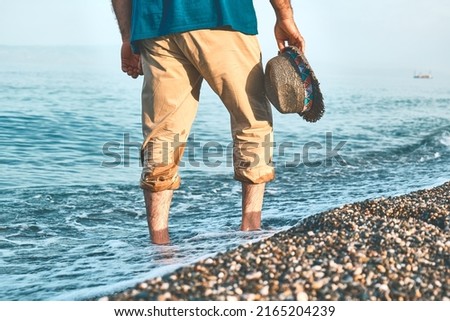Man wearing shorts, walking barefoot along the seashore. Male legs walks on pebble beach along the shore near the water with waves, low section. Wellness, freedom and travel in summertime concept. Royalty-Free Stock Photo #2165204239