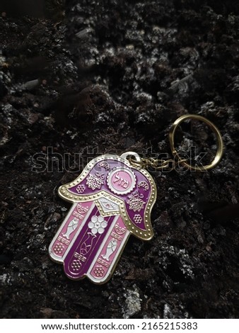 Pink-purple metal keychain Jewish "Hand of Miriam" on a black ground background close-up with the inscription "Good luck". 
Hamsa is the popular protection amulet in the Middle East and North Africa.