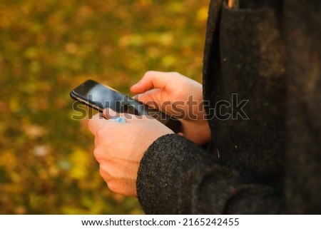 Defocus male hand holding phone. Man using smart phone during resting in autumn park. Typing text message or reading social media at mobile phone. Fall background. Out of focus.