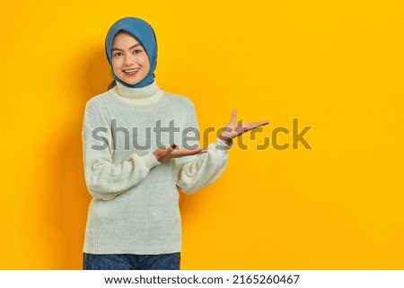 Cheerful beautiful Asian woman in white sweater presenting copy space on palm isolated over yellow background
