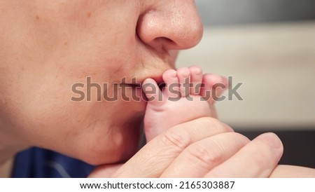 Grandma kisses bare tiny feet with little fingers of newborn baby on blurred background. Woman shows love for little granddaughter in bedroom closeup