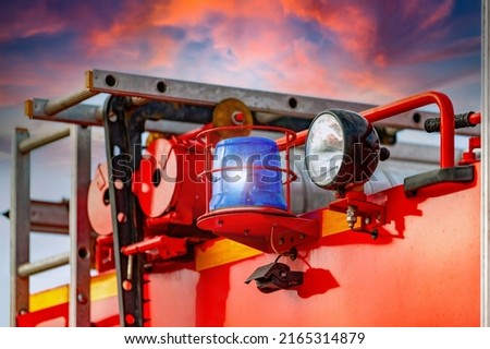 Close-up of a blue flashing light of a siren on a red car of firefighters, ambulance or rescuers against the background of a burning sky - a fire reflected in the clouds.