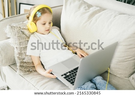 Little girl 4 years old in yellow headphones in front of a laptop. The child looks at the computer screen. The baby watches cartoons while mom is busy. Remote learning for preschool children.