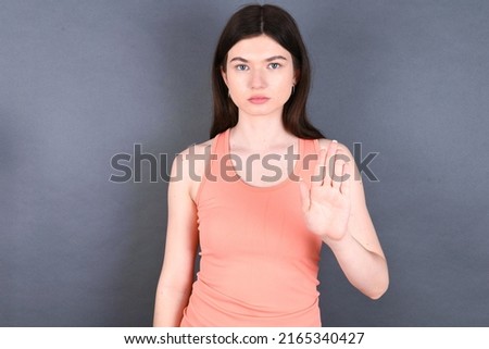 young beautiful Caucasian woman wearing orange shirt over studio grey wall shows stop sign prohibition symbol keeps palm forward to camera with strict expression