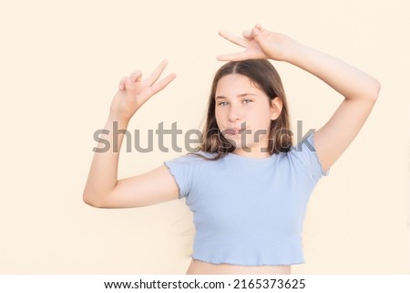 Young Caucasian girl making funny gesture with tongue out and fingers in victory sign on beige isolated background