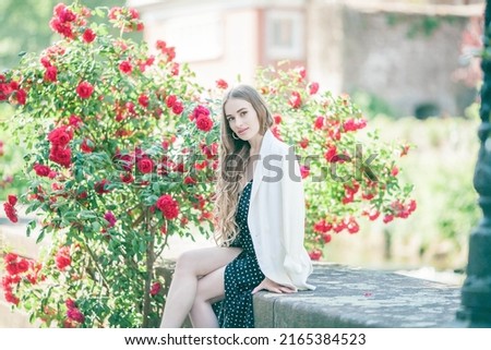 Portrait of a young beautiful slim long-haired woman in a romantic black and white dress near a red blooming rose in the center of a European city. Model 23 years old in classic clothes.