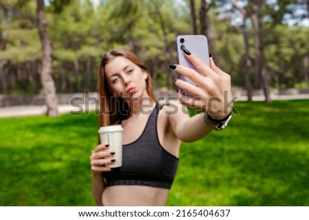 Happy brunette woman wearing black sports bra standing on city park, outdoors taking selfie and holding takeaway coffee mug. Self portrait for social media. Outdoor sport concepts.