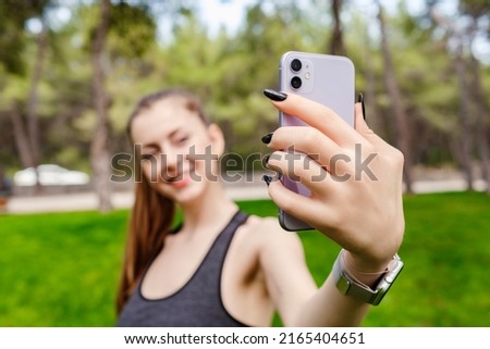 Cute caucasian woman wearing sportive clothes on city park, outdoors taking a self portrait with smart phone. She looks at the screen and taking selfie. Selective focus on phone.
