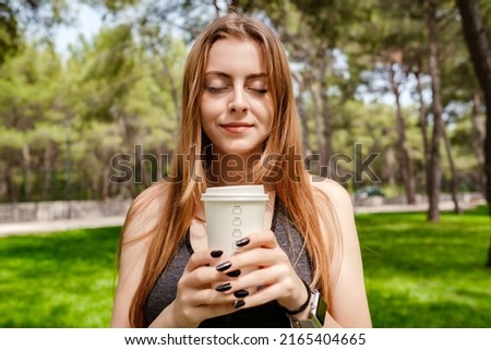Portrait of young brunette woman wearing sportive clothes on city park, outdoors holding a takeaway coffee mug front of chest and closed eyes. Coffee lover and outdoor sport concepts.