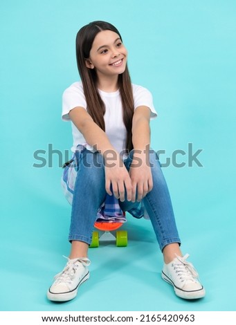 Happy vogue teenager. Fashionable confident teenage girl wearing trendy t-shirt and jeans sit on skateboard, posing on blue background. Fashion teenager, casual look.