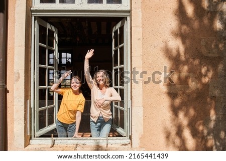 Cheerful woman and her daughter waving to somebody while looking from the window Royalty-Free Stock Photo #2165441439