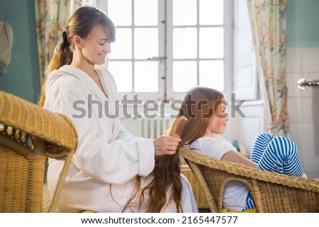 Friendly woman combing hair of her daughter at home