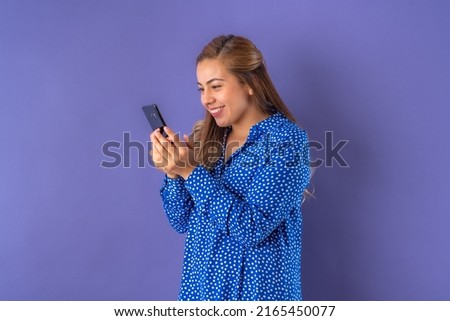 Beautiful attractive young woman posing happily while holding mobile phone, in blue dress with dots. Isolated over purple background.