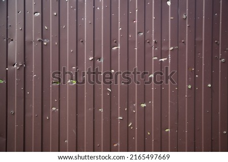 Textured background of a brown metal fence that has been shelled. The surface is covered by gunshot residue. The shelling of the building. Vertical lines on a flat plane. War in Ukraine 2022. Royalty-Free Stock Photo #2165497669