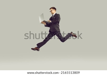 Competition in business. Successful young businessman who is in hurry to do lot of work working on laptop on run. Man in suit and glasses looking to enhance his career is isolated on gray background.