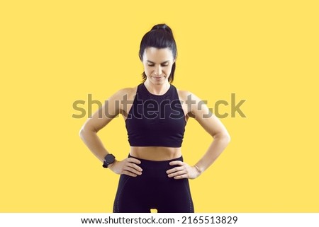 Confident sportswoman holding hands on waist standing in workout pose on vivid yellow background. Woman with fit body in black sportsbra, leggings and with smart watch performs physical exercises.