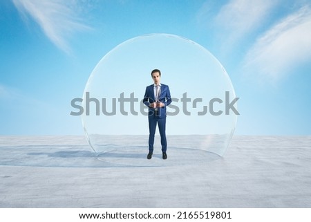 Attractive young european businessman standing under and inside big glass sphere cover on concrete ground and bright blue sky background. Protection and limitation concept
