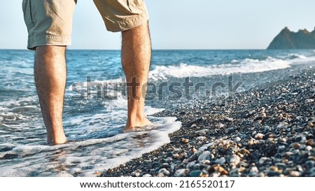Man wearing shorts, walking barefoot along the seashore. Male legs walks on pebble beach along the shore near the water with waves, low section. Wellness, freedom and travel in summertime concept. Royalty-Free Stock Photo #2165520117