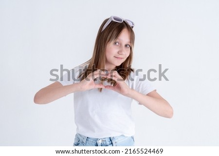 Stylish teen girl with long blond hair and blue eyes in a white t-shirt and blue jeans shows a heart shape with her fingers and smiles on a white background. Space for text studio photo.