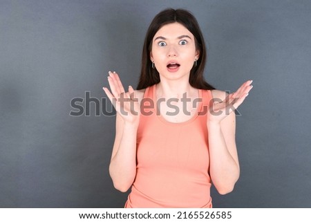 Surprised terrified Caucasian woman wearing orange sportswear over studio grey wall Gestures with uncertainty, stares at camera, puzzled as doesn't know answer on tricky question, People, body languag