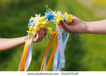 Close-up on bright Ukrainian wreath with yellow and blue wild flowers and long multi-colored ribbons in hands of unknown Ukrainian teenage children, that brings hope for peaceful sky and end of war