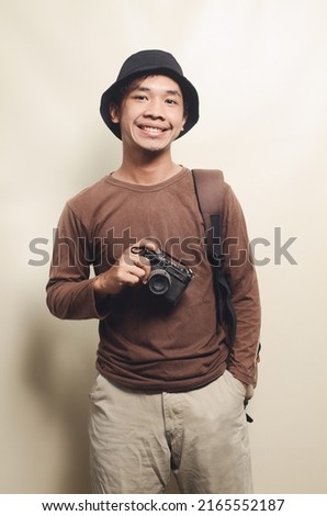 Portrait of cheerful young Asian man wearing black hat carrying camera and backpack isolated on background