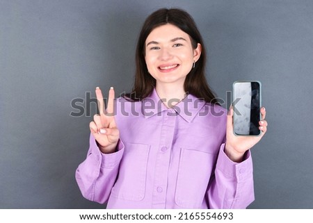 young caucasian woman wearing purple shirt over grey background holding modern device showing v-sign