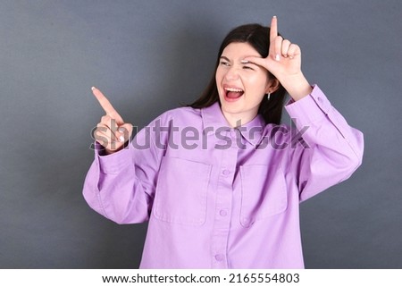 young caucasian woman wearing purple shirt over grey background showing loser sign and pointing at empty space