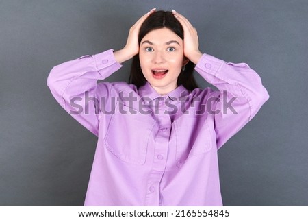 Cheerful overjoyed young caucasian woman wearing purple shirt over grey background reacts rising hands over head after receiving great news.