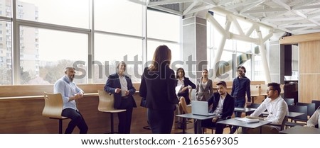 Various businessmen listen to female business leader during corporate meeting or training lecture. Rear view of woman in business attire speaking to people in modern business center. Panoramic view. Royalty-Free Stock Photo #2165569305