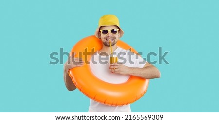 Happy young man with inflatable swimming circle and with glass of juice enjoys summer vacation. Cheerful funny guy in summer clothes drinks juice on light blue background. Summer relaxation concept. Royalty-Free Stock Photo #2165569309